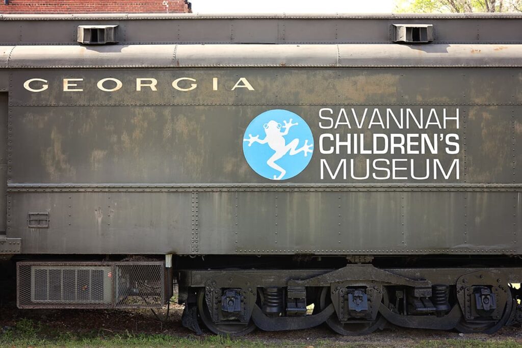 Faded black railcar with the words" Georgia" and "Savannah Children's Museum" in white text and a white tree frog on a pale blue circular background