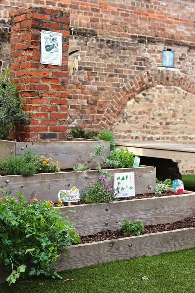 Four level wooden planter filled with plants and colorful markers. An old brick wall is visible in the background