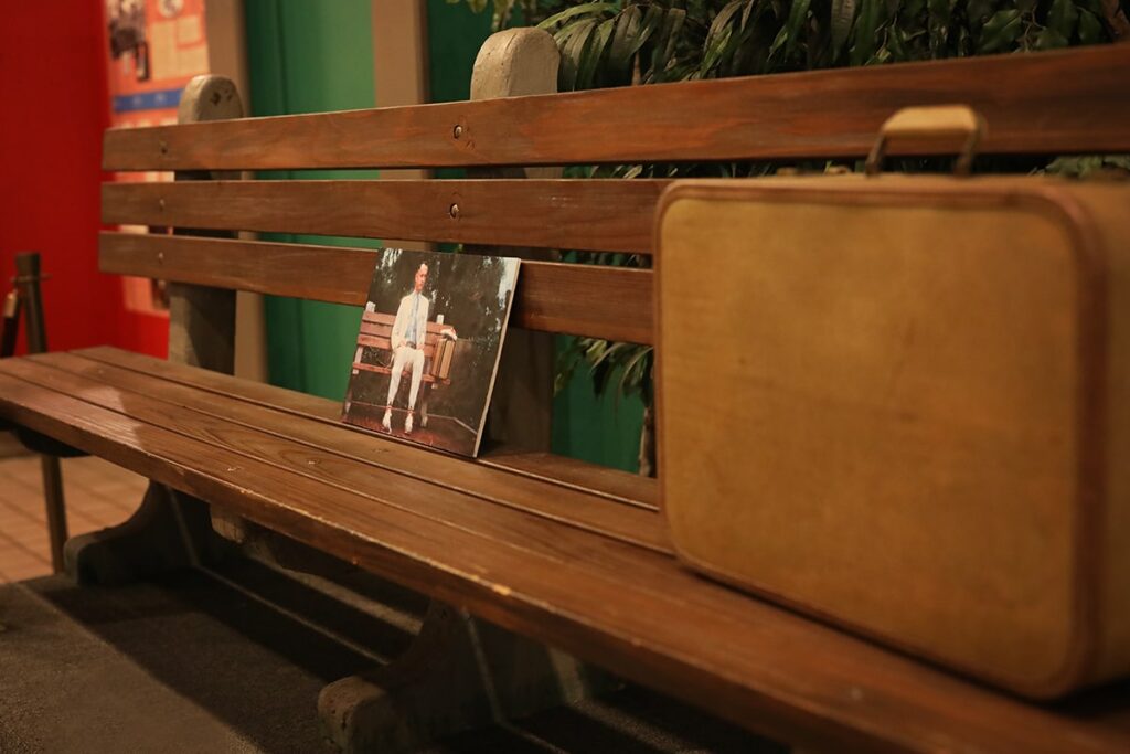 A photo of Forrest Gump sitting on a bench in Chippewa Square is leaning against the brown wooden slats of the actual bench where he sat. His suitcase is located next to the photo