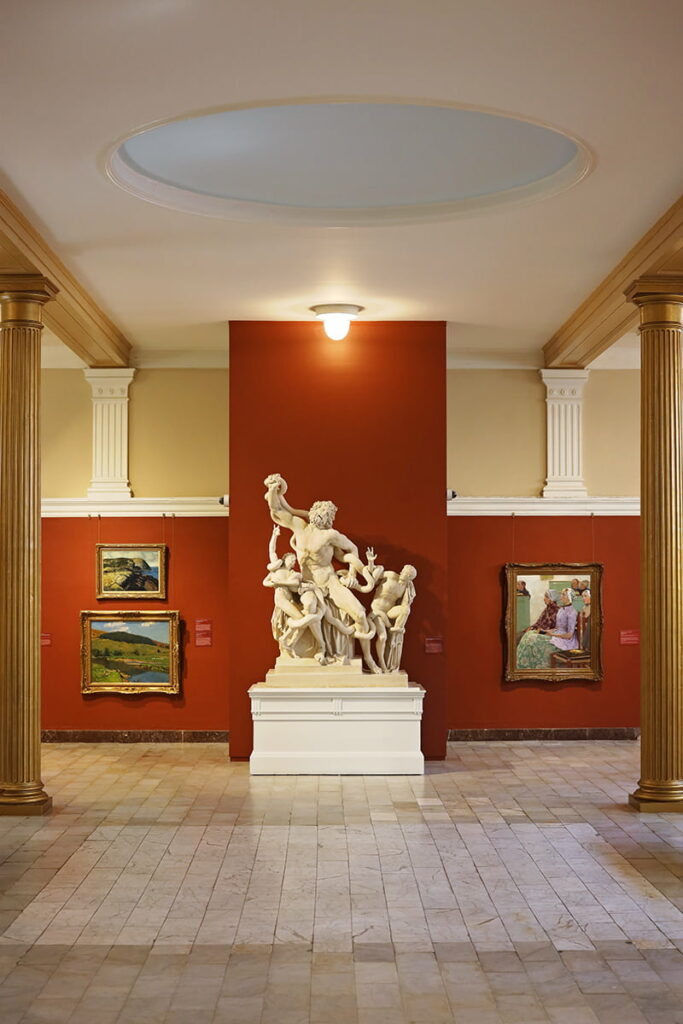 Marble statue framed against a deep-red painted wall in Telfair Academy with elegant gold columns and gold-framed art on each side