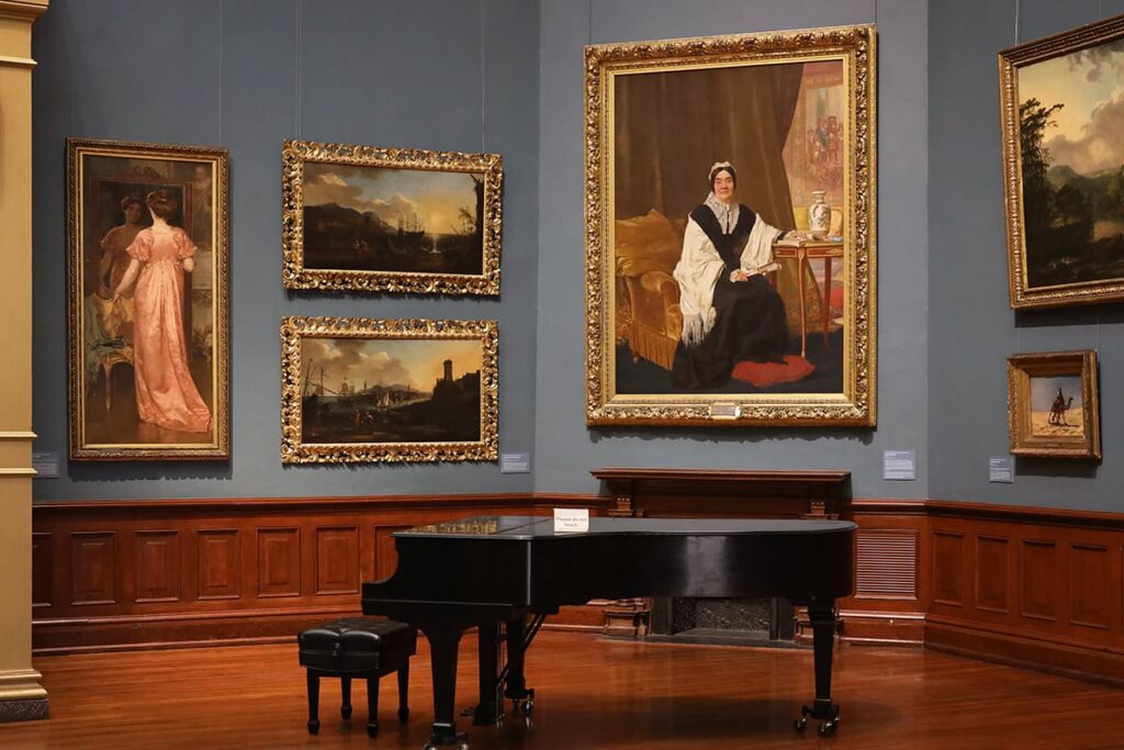 Interior of the Telfair Academy showing oversized art in gold frames against a greyish-blue wall with elegant wooden trim. A piano sits in the foreground