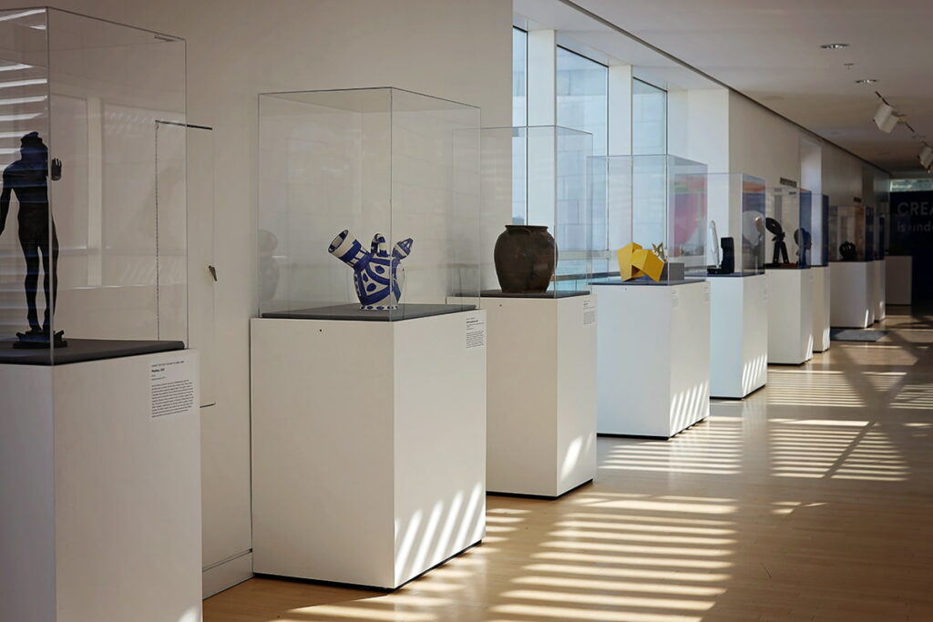 Rows of pottery and sculptures framed in glass boxes against a wall inside the Jepson Center. The sun's rays are reflecting light on the ground in unique striped patterns