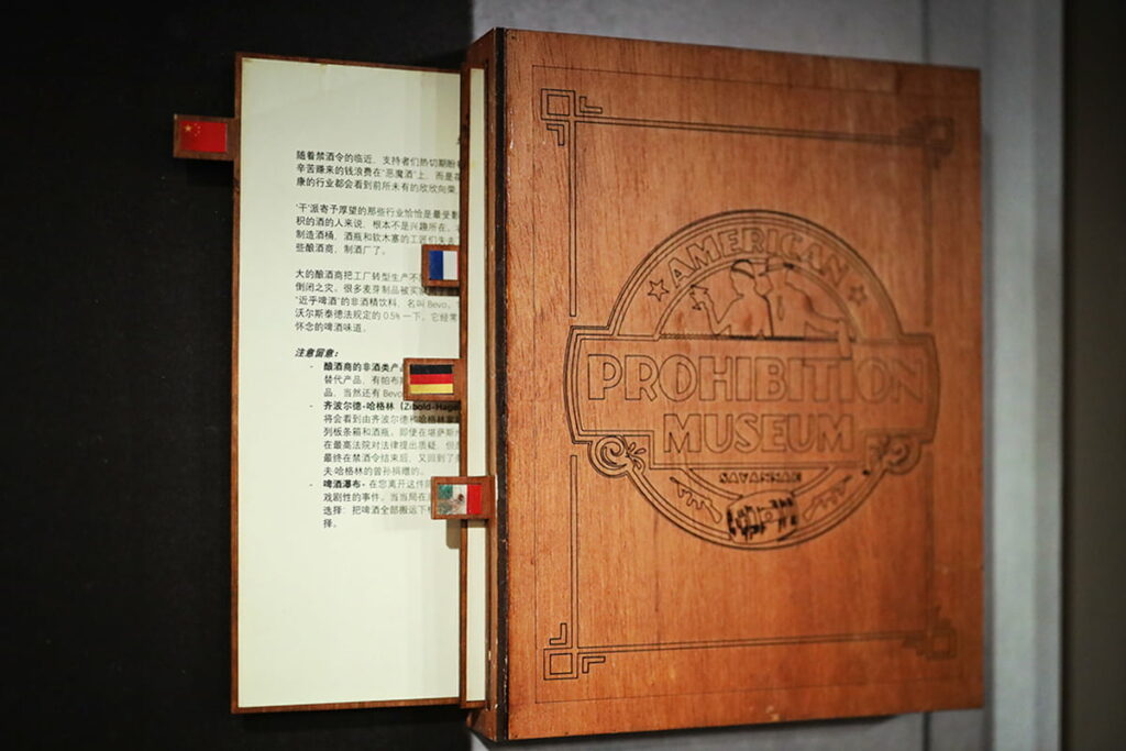 Wooden box mounted on the wall with the American Prohibition Museum logo etched into the front cover. Pullout tabs represent Chinese, French, German, and Mexican flags