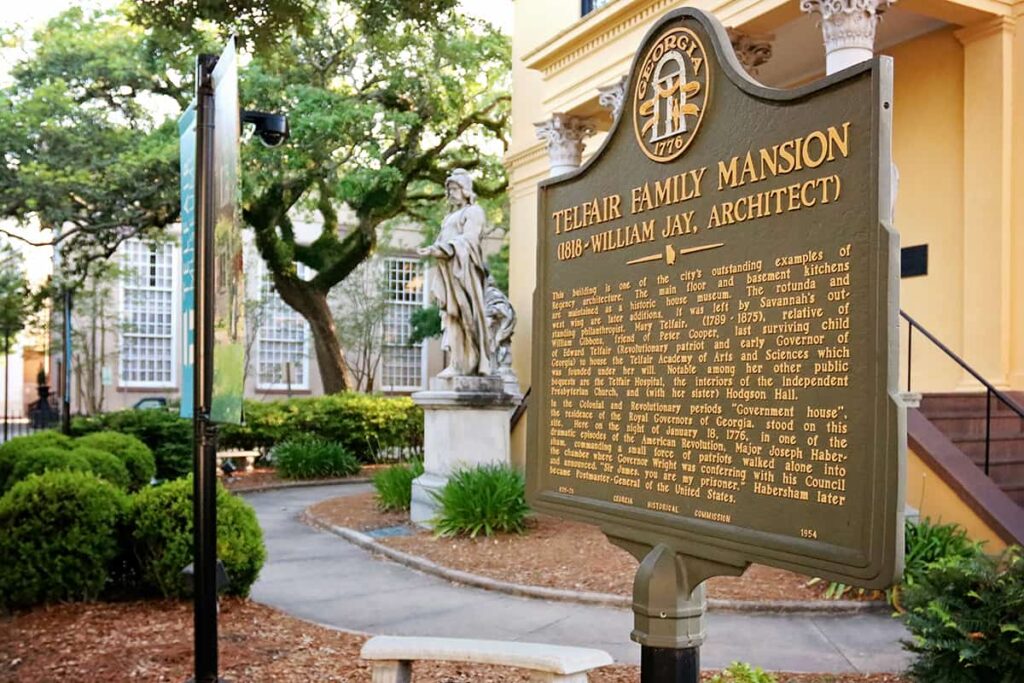 Georgia Historic Marker for the Telfair Family Mansion (1818 - William Jay Architect) stands in front of the yellow-painted stucco façade of Telfair Academy. Large oaks and a few statues are visible in the courtyard in front of the building