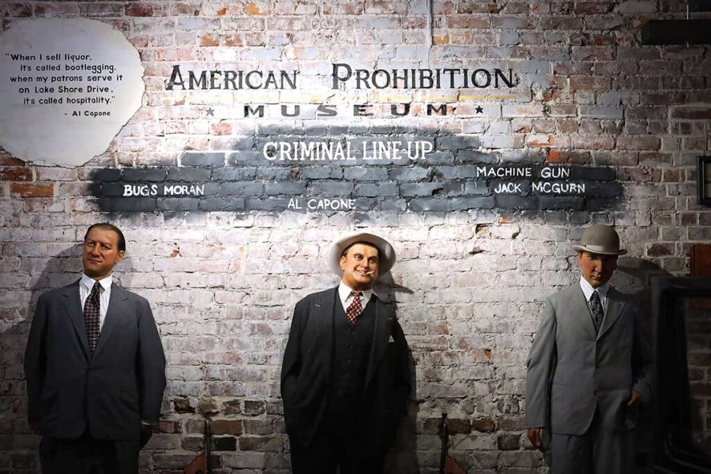 A criminal lineup against a brick wall inside American Prohibition Museum shows wax figures for Bugs Moran, Al Capone, and Machine Gun Jack McGurn. All three are dressed in dapper gray suits