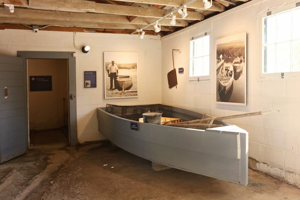 An old wooden crabbing rowboat painted in a pale shade of blue is on display beneath B&W photos of members of the Pin Point community at Pin Point Heritage Museum in Savannah
