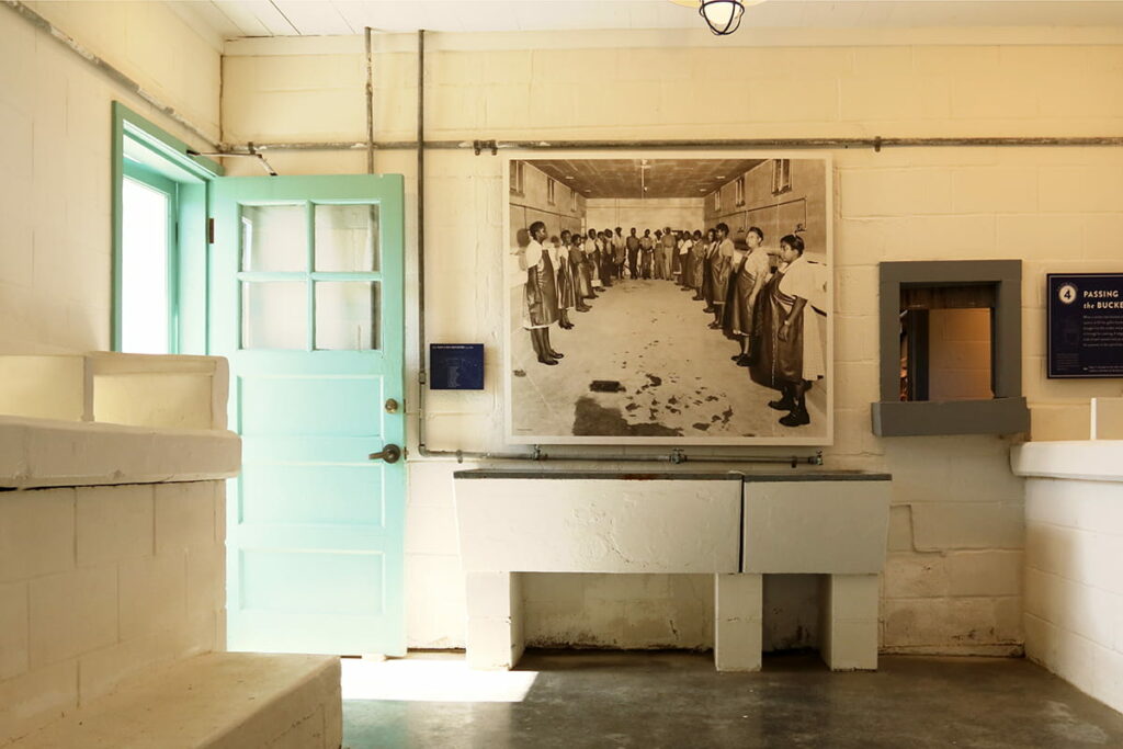 A room with concrete walls and floors and numerous oyster-shelling stations at Pin Point Heritage Site. Historic B&W photos of former employees line the walls