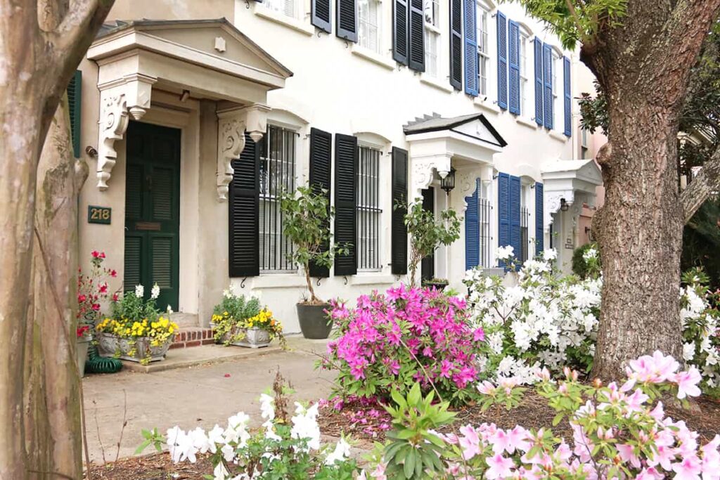Off-white rowhomes in Savannah with black shutters on one and blue shutters on the other, with an assortment of pink and white azalea bushes in the garden