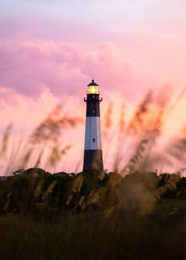 Peering through grassy beach dunes towards the Tybee Island lighthouse with a beautiful peach and pink sunset in the background