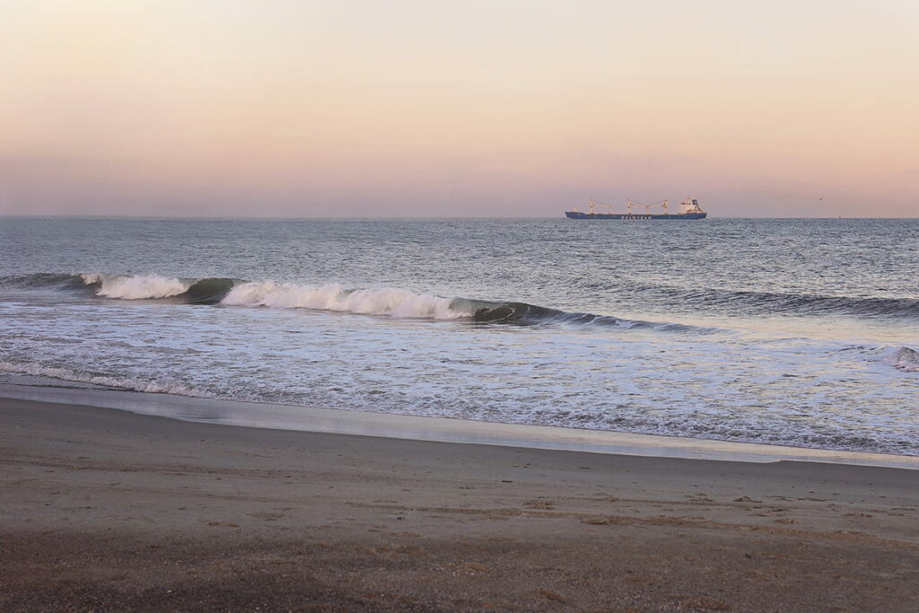 Crashing waves along North Beach in Tybee Island Georgia with a pale pink sunset and a cargo ship visible on the horizon