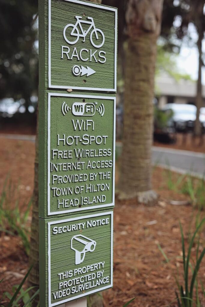 Three square green wooden signs with white lettering. The first sign indicates the direction of bike racks, while the second sign announces the availability of free WiFi. The third sign reads "Security Notice: This property is protected by video surveillance"