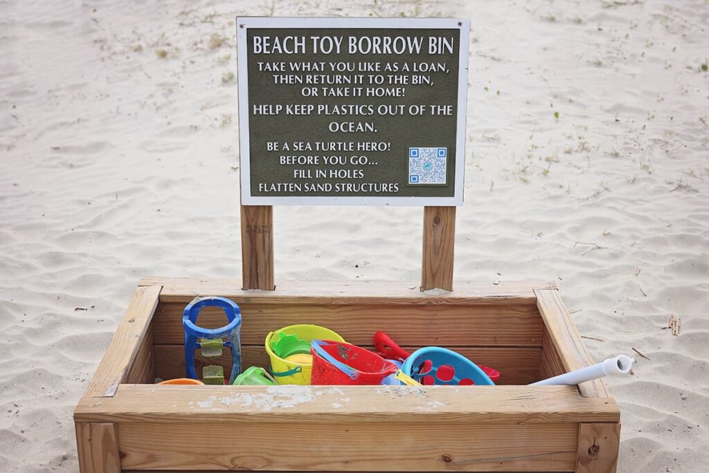 A wooden box filled with colorful toys sits on Coligny Beach with a green wooden sign indicating it is a "Beach Toy Borrow Bin"