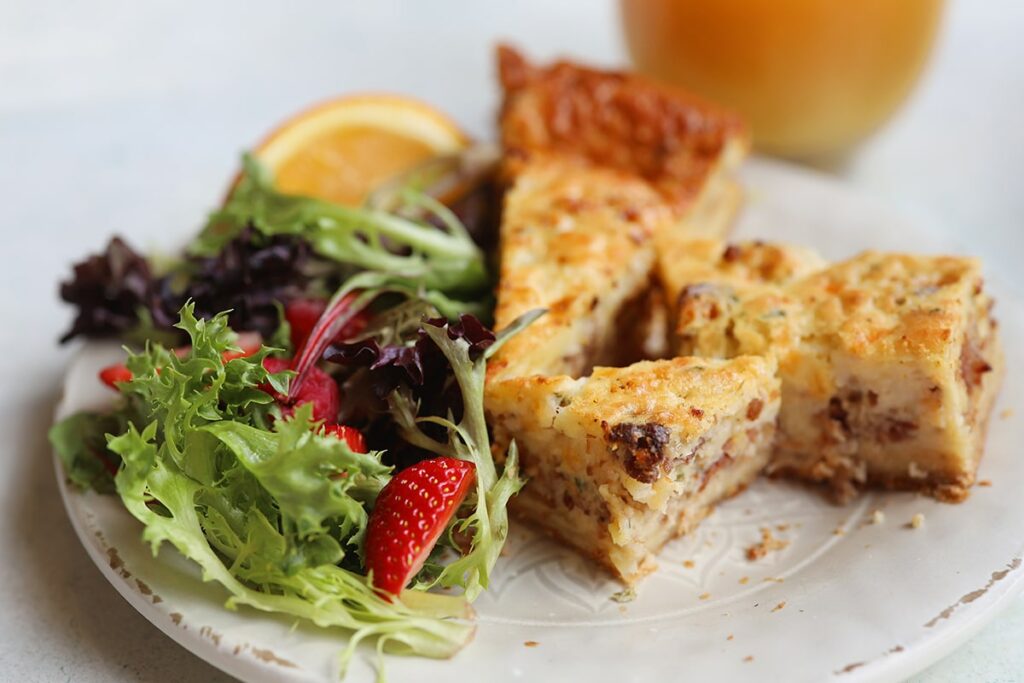 Three slices of a thick and flaky quiche sit on an off-white plate at Cafe M in Savannah surrounded by a fresh salad of greens, strawberries, and orange slices