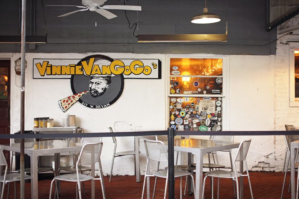 Covered patio with ceiling fans and numerous groupings of white metal tables and chairs. The Vinnie Van Go Go's label and an image of a chef with a slice of pizza is visible against a white wall in the background