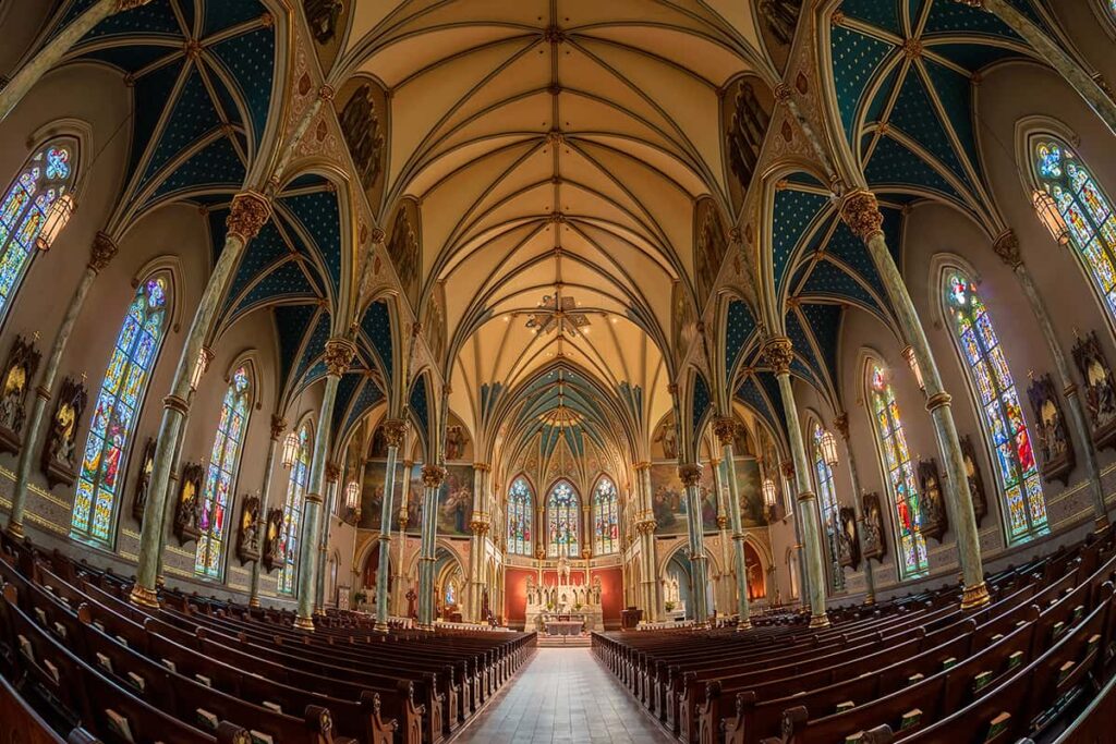 A wide angle shot of the interior of the Cathedral Basilica of St. John the Baptist. It appears cavernous, with high ceilings, endless rows of pews, and numerous stained glass windows that are each approximately 20 feet tall