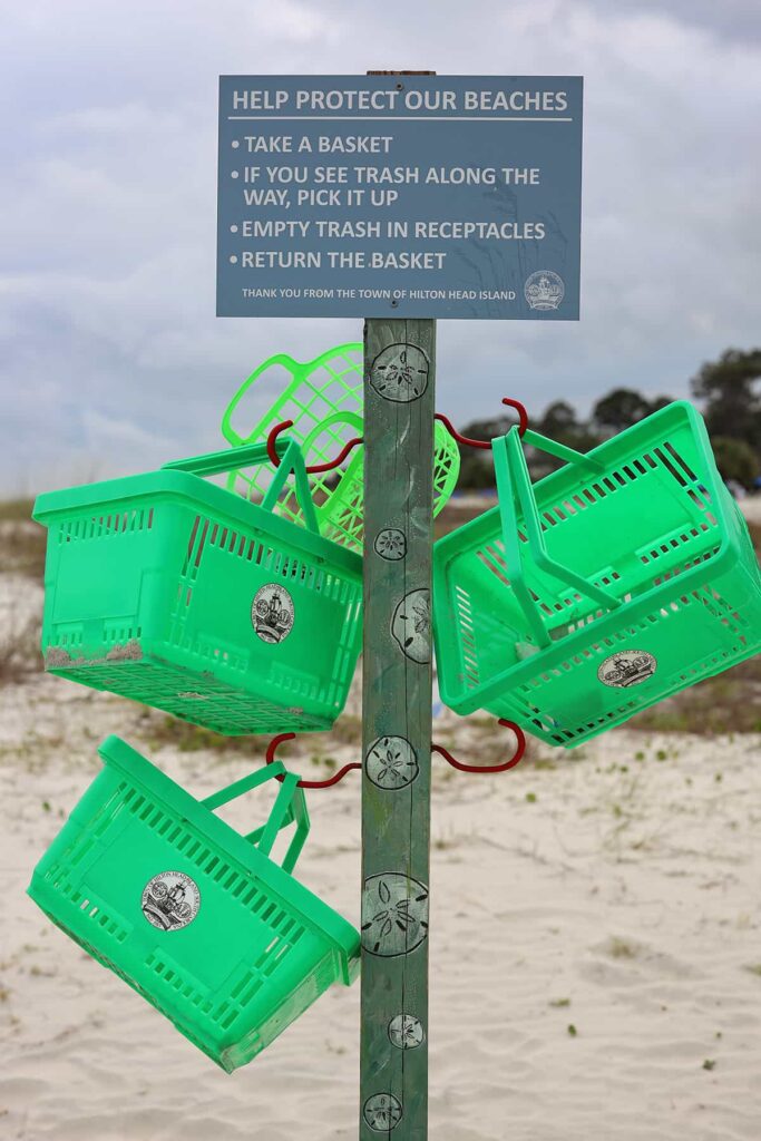 Wooden post with green baskets hanging from it on Coligny Beach. An attached sign reads: "Help Protect our Beaches: Take a basket. If you see trash along the way, pick it up. Empty trash in receptacles."