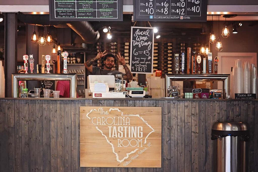 A friendly tattooed employee smiles and flashes two peace signs from behind the counter at The Carolina Tasting Room in Coligny Plaza