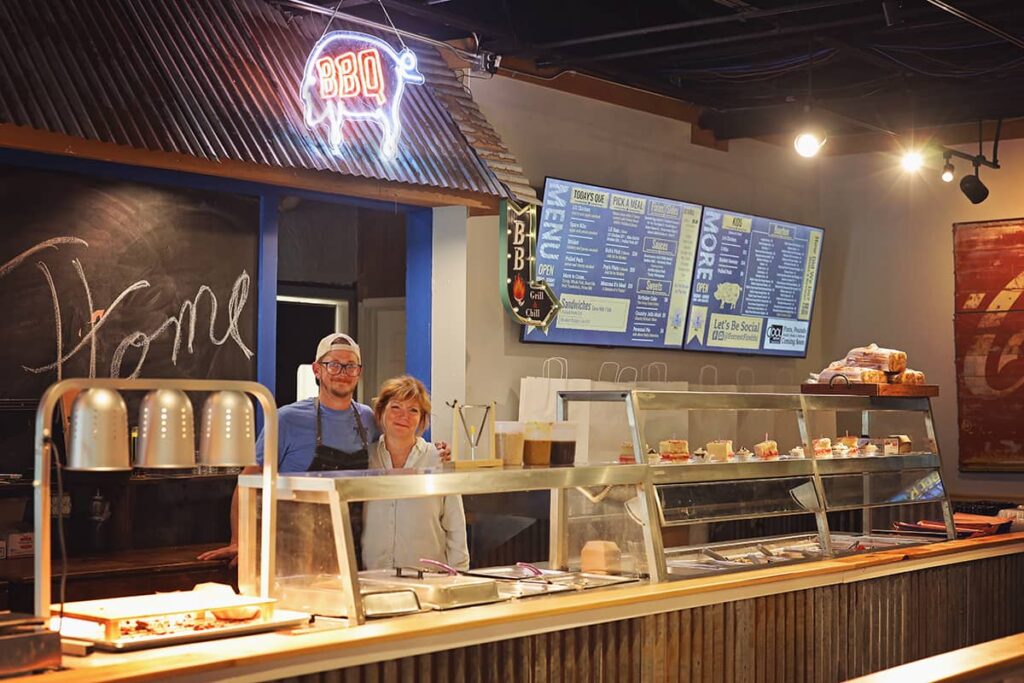 The owners of Forest Fire BBQ, a young male wearing a backwards ball cap and a female with a friendly smile, pose behind the restaurant's counter. A neon sign hangs in the background with the outline of a pig and the word "BBQ" lit up in yellow and red