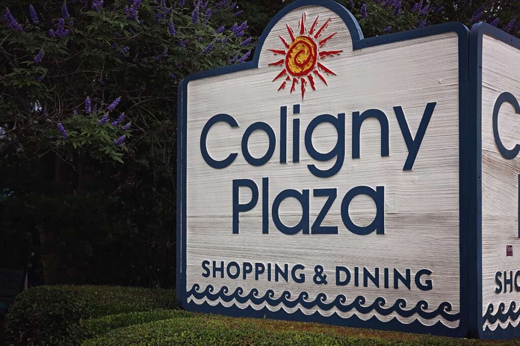 The Coligny Plaza sign in light grey driftwood with a navy blue outline and navy blue lettering. Purple Mountain Laurel blooms are visible on a tree in the background