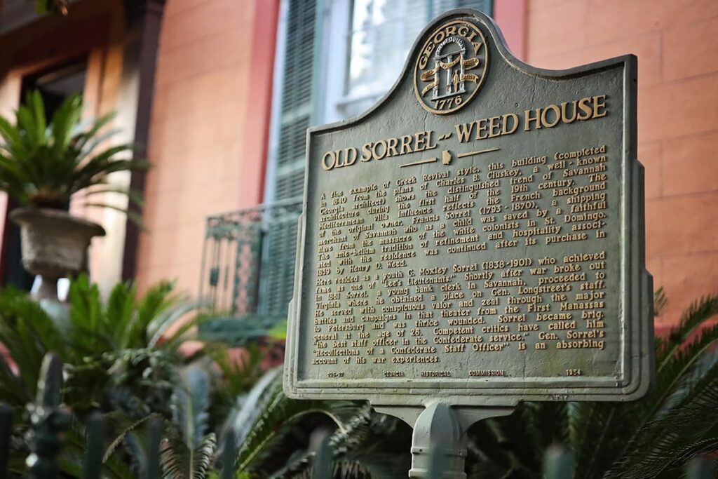 Historic Marker for the Old Sorrel-Weed House, one of the haunted houses in Savannah, with green sago palms in the foreground and an orange house with green shutters in the background