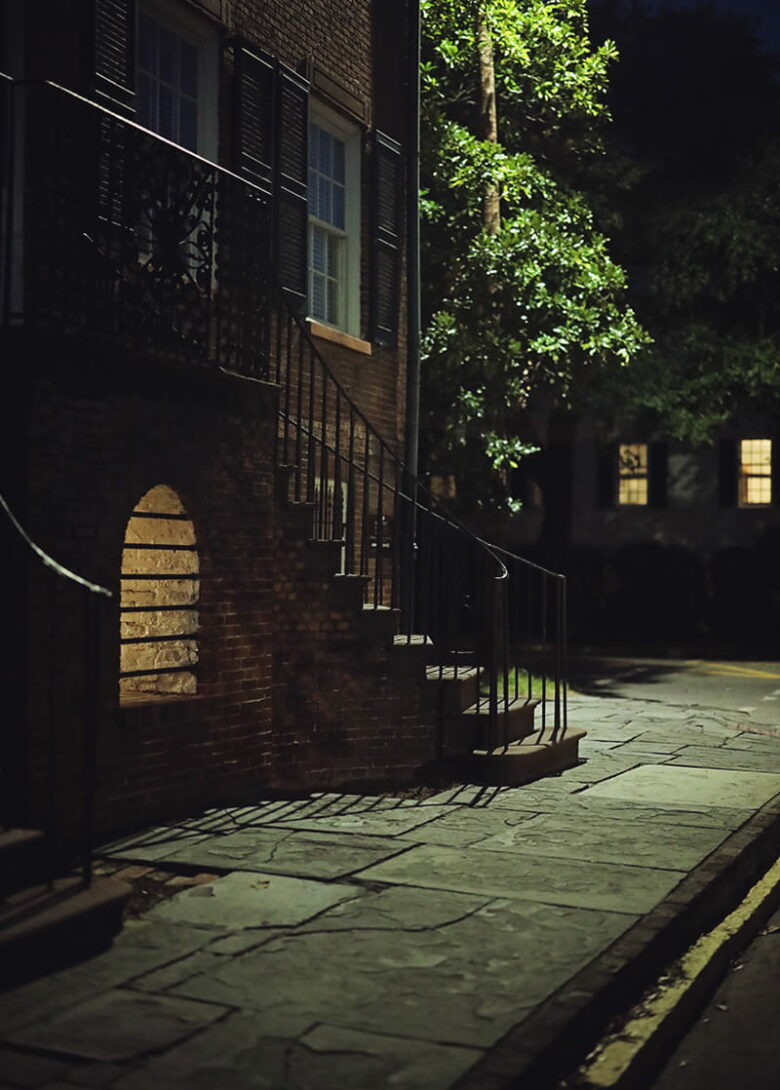 The façade of the Davenport House in Savannah looks spooky at night, with a pale yellow glow emanating from a barred vault located between the home's curved front staircases