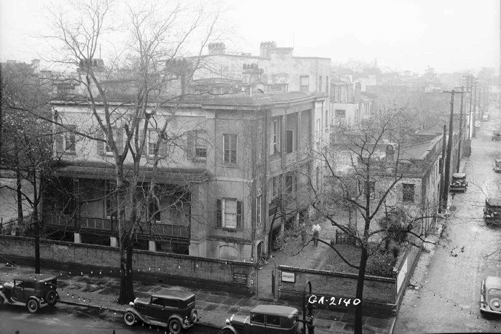 Historic B&W aerial view of the three-story Sorrel-Weed House surrounded by a brick wall. The surrounding trees are bare and the streets are wet as if it rained recently. Old Model T Ford-style vehicles are parked in the road out front