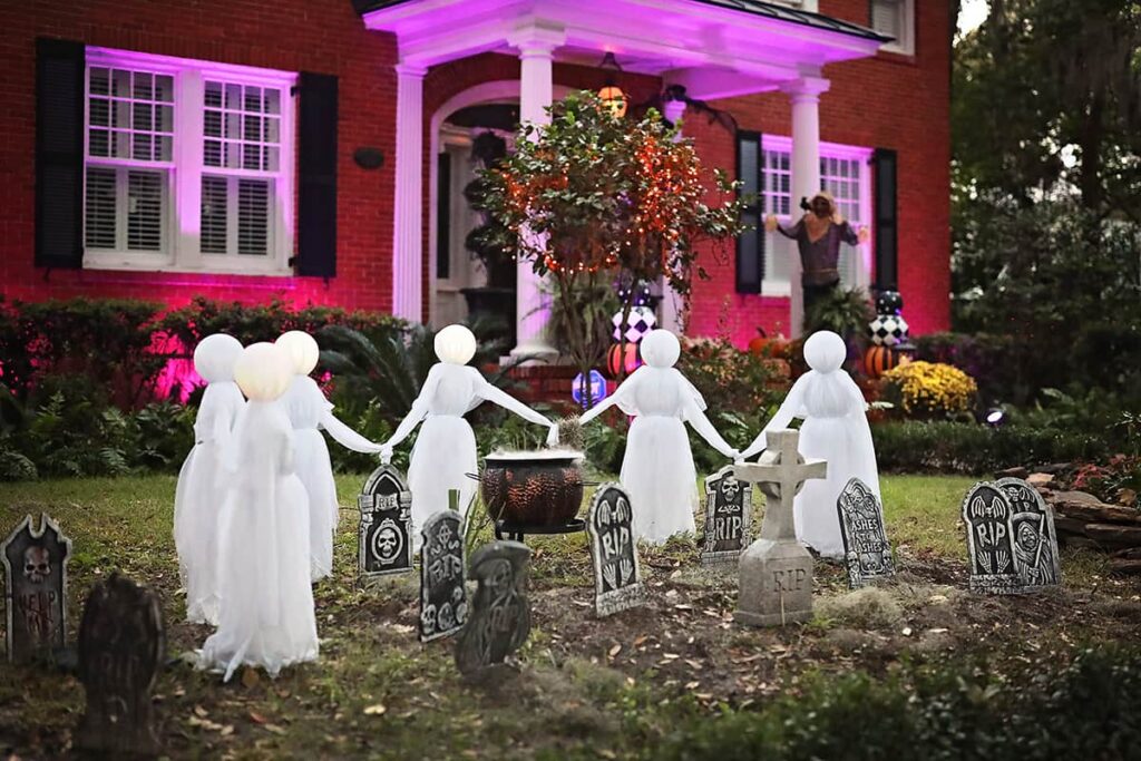 A coven of witches dressed in white hold hands and form a partial circle around headstones in the front yard of a stately brick home in Ardsley Park, Savannah
