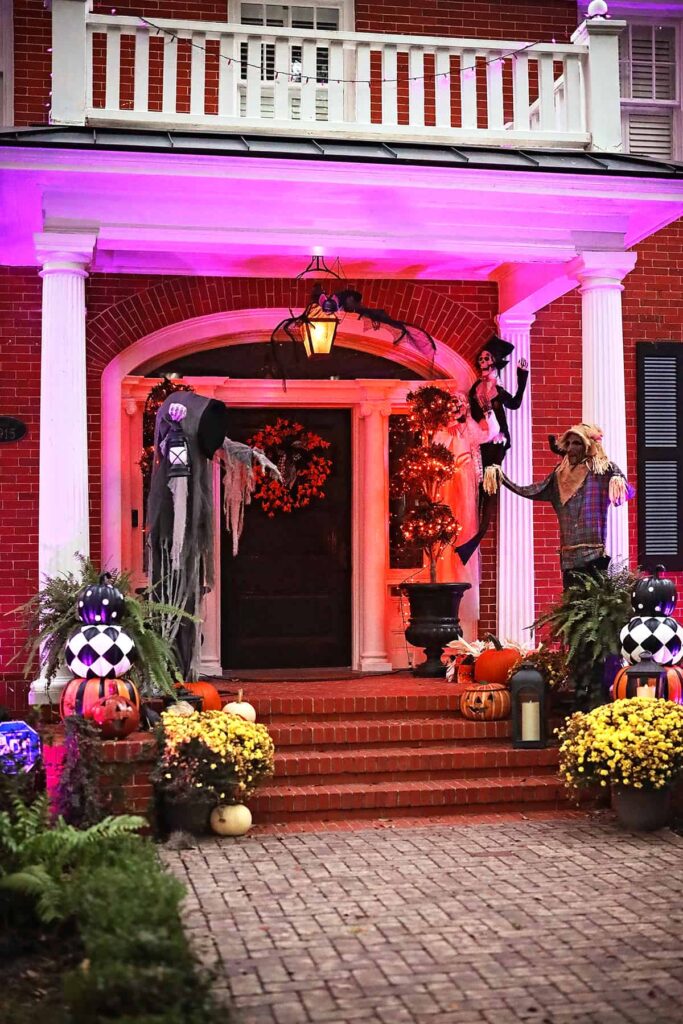 The front porch of a stately brick home is decorated for Halloween in Savannah with yellow mums, pumpkins, a scarecrow, giant spiders, and additional spooky creatures