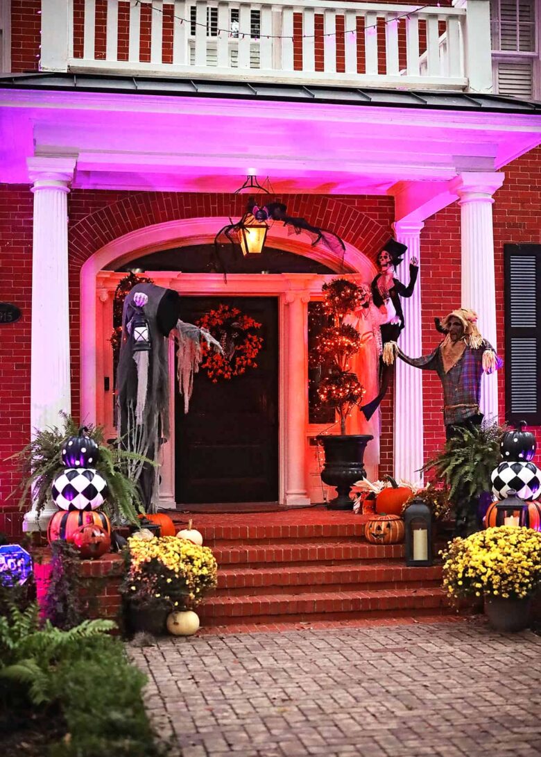 The front porch of a stately brick home is decorated for Halloween in Savannah with yellow mums, pumpkins, a scarecrow, giant spiders, and additional spooky creatures