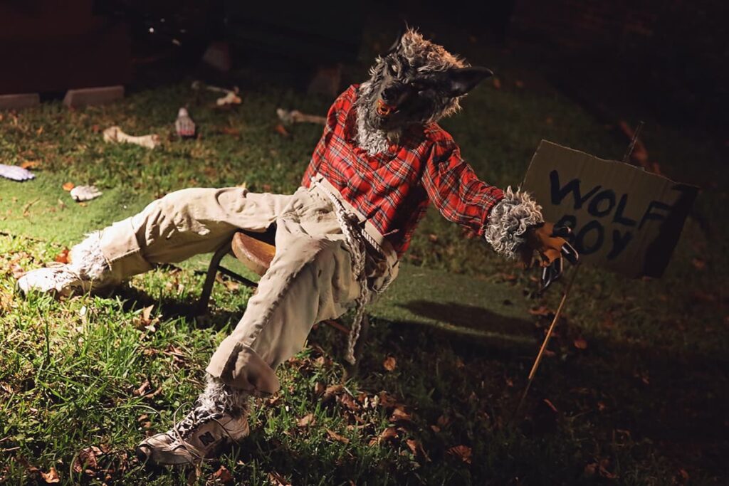 A figure in baggy tan pants and a red plaid shirt rests on a stool with a wolf mask covering his face and tufts of fur peeking out from beneath his clothing. A nearby cardboard sign has the words "wolf boy" written in crude black lettering