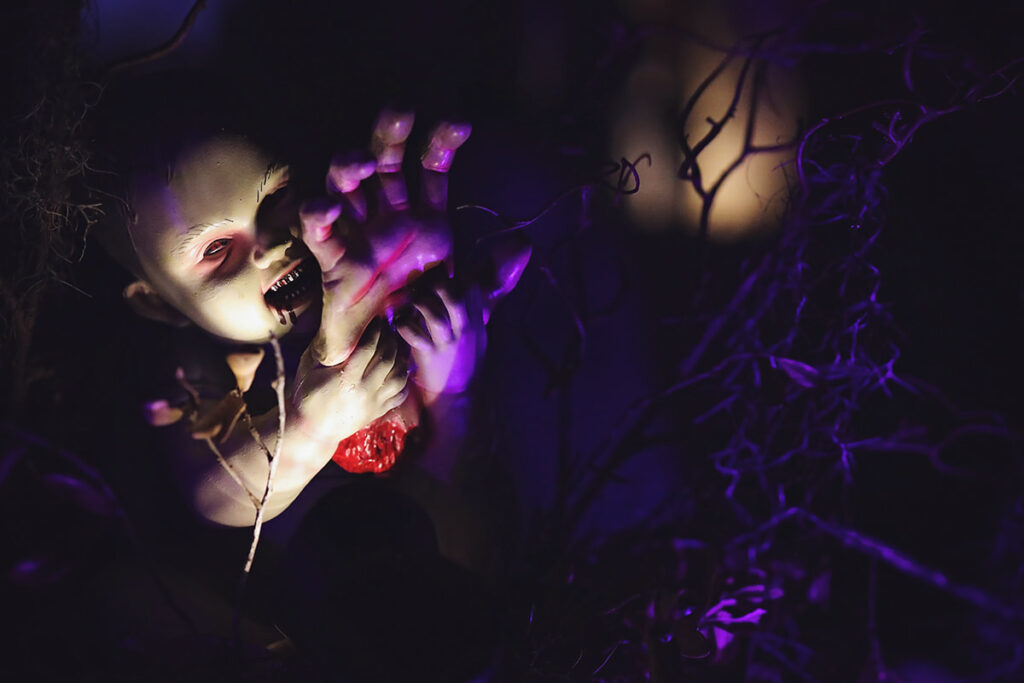 Purple lighting illuminates a creepy doll with red eyes and jagged teeth. The doll is holding a severed hand to its mouth, as if about to take a bite