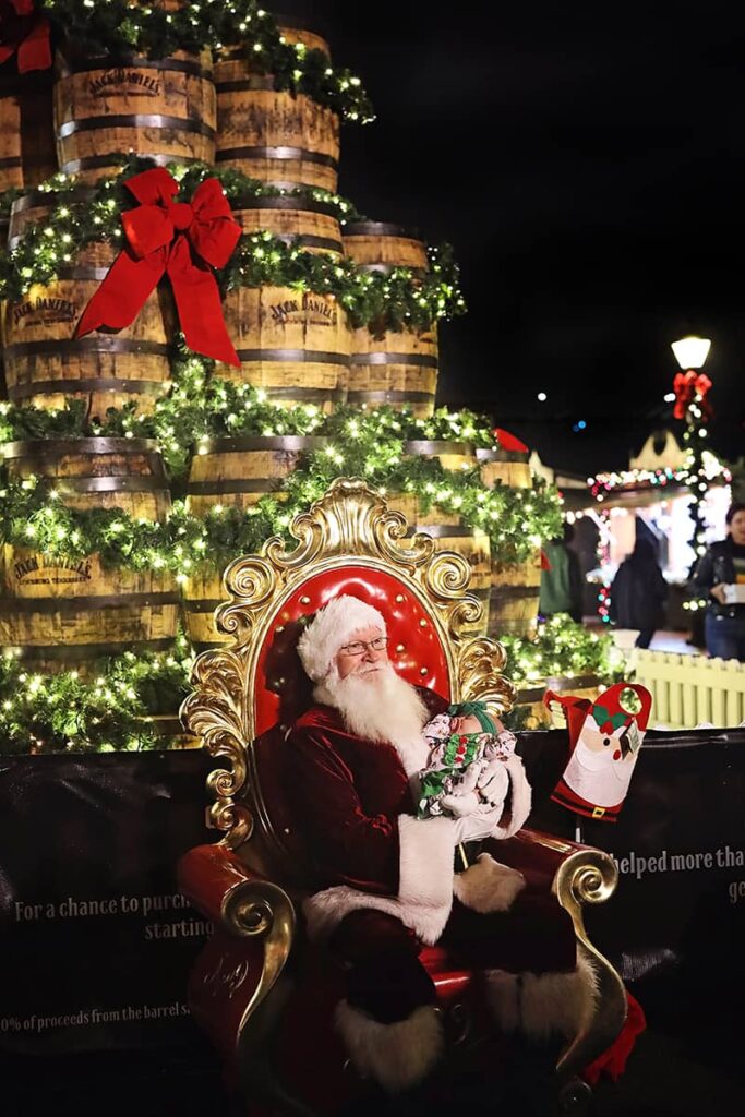 Santa Clause holds a baby while sitting in a red and gold chair in front of holiday decor at the Savannah Christmas Market