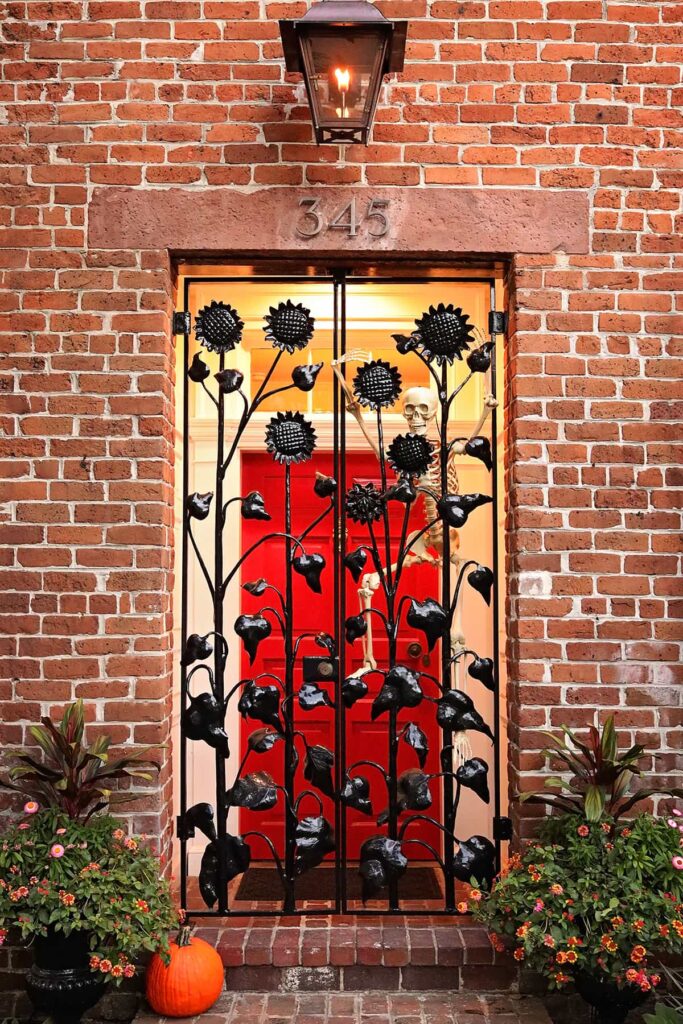 A stately brick home features a red front door and decorative outer doors made of wrought-iron sunflowers. The home is decorated for Halloween in Savannah with a skeleton peering out from behind the sunflowers as if to say, "set me free"