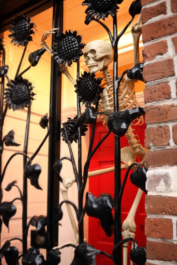 A skeleton peers out from behind a decorative door made of black wrought-iron sunflowers