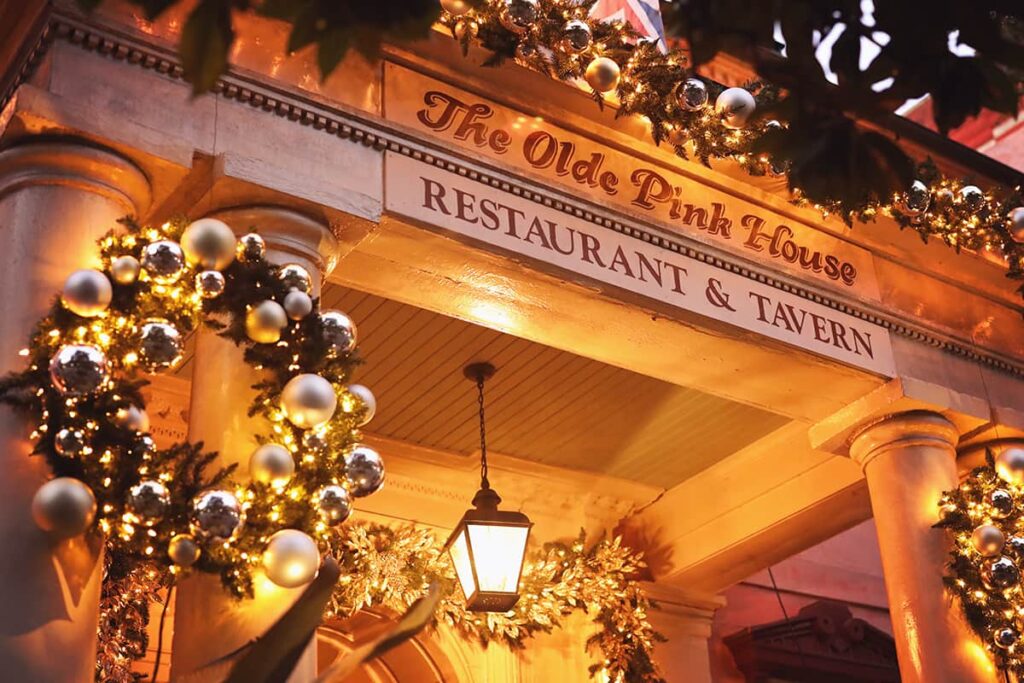 A painted wooden sign with the words "The Olde Pink House Restaurant & Tavern" hangs above a front porch entryway decorated by garlands and wreaths covered in silver ornaments