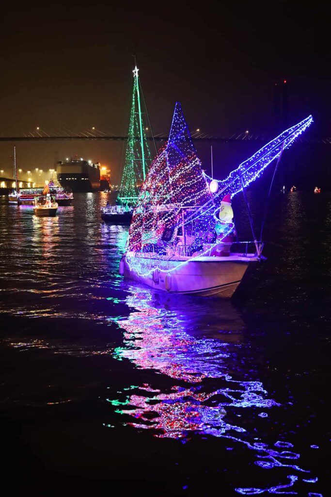 A lighted parade of boats in floats down the Savannah river with the Talmadge bridge and a large container ship visible in the background