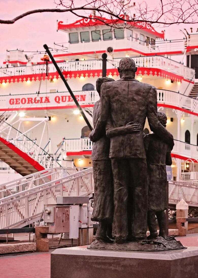 View of the African American Monument on River Street with the Georgia Queen Riverboat illuminated by a pale pink sunset visible in the background