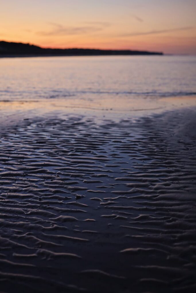 Wavy layers of pluff mud on Mitchelville Beach in the foreground with hues of a yellow and lavender sunset reflected in the ocean in the background
