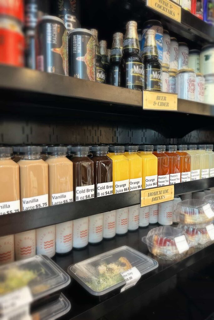 Bodega-style shelves in Dottie's Market with grab-and-go sections for homemade drinks, beer and cider, wine and cocktails, and packaged foods such as quiche and desserts