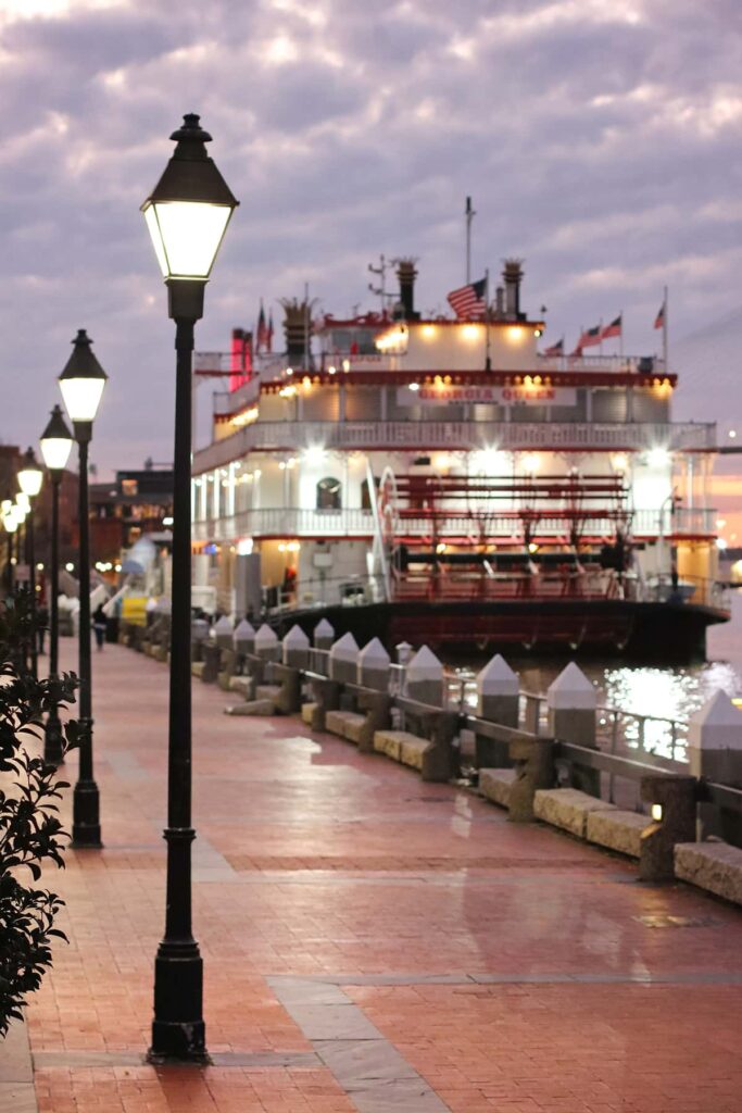 The Georgia Queen Riverboat docked on River Street with gas lanterns visible on Rousakis Plaza in the foreground and a cloudy lavender sunset in the background