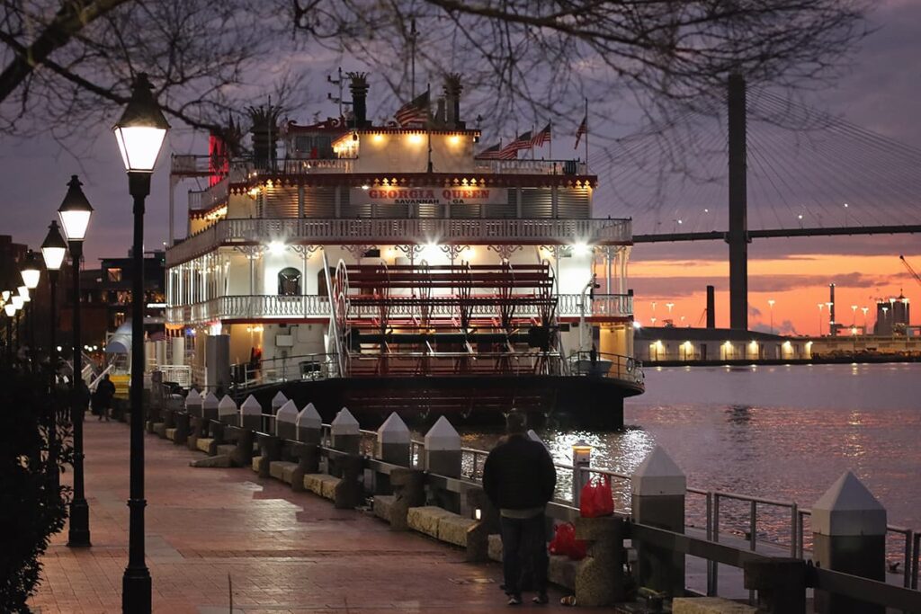 The Georgia Queen Riverboat docked along River Street at night with a lavender and orange sunset visible behind the Talmadge Bridge