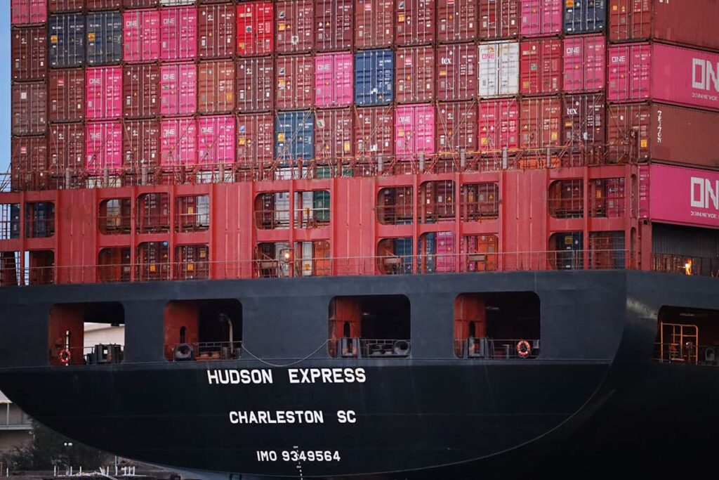 The stern of the Hudson Express based out of Charleston SC shows dozens of pink, red, blue, white, and green containers stacked atop its deck as it heads out to sea