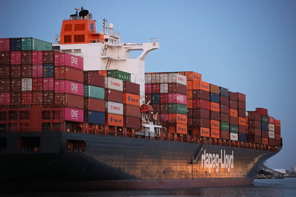 The Hudson Express cargo ship leaves the port of Savannah with colorful containers stacked atop its deck. The words "Hapag-Lloyd" appear on the starboard side in blocky white lettering