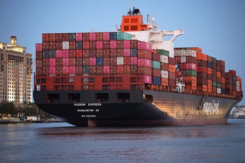 The Hudson Express cargo ship leaves the port of Savannah with colorful containers stacked atop its deck. With so many containers stacked aboard, the ship appears as tall or taller than a many-multi-story hotel that can be seen nearby along the banks of the river