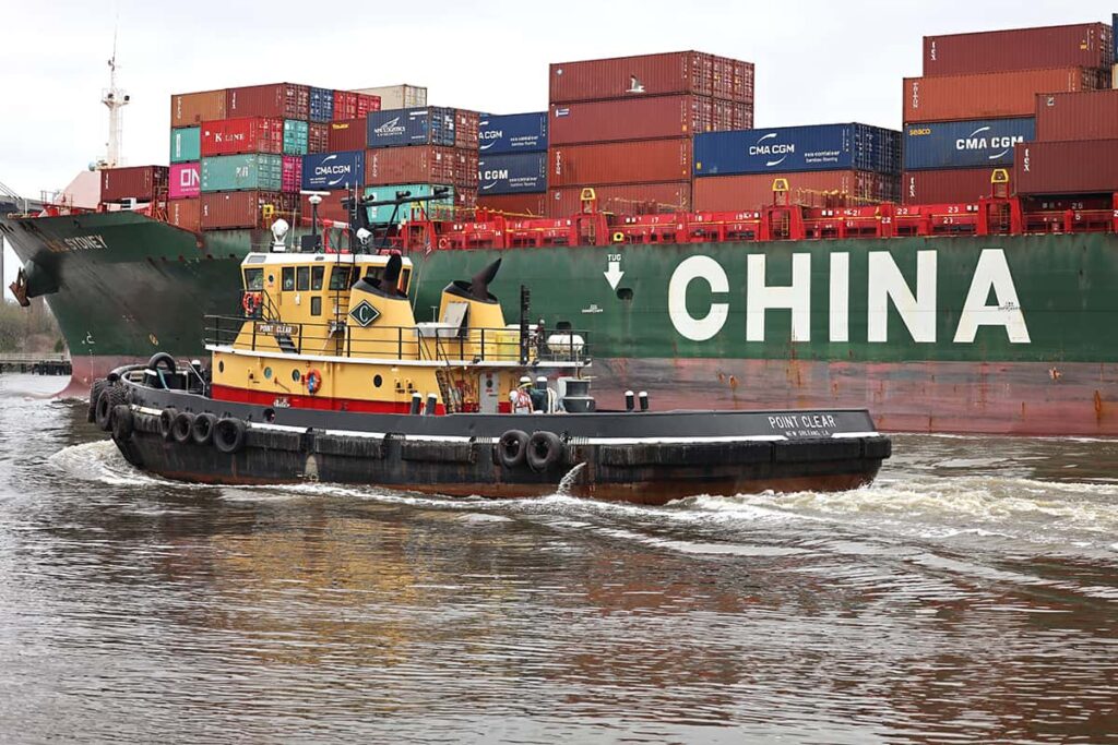 A yellow, red, and black tugboat named POINT CLEAR cruises alongside of a large green cargo ship with the word CHINA labeled in white across its port side