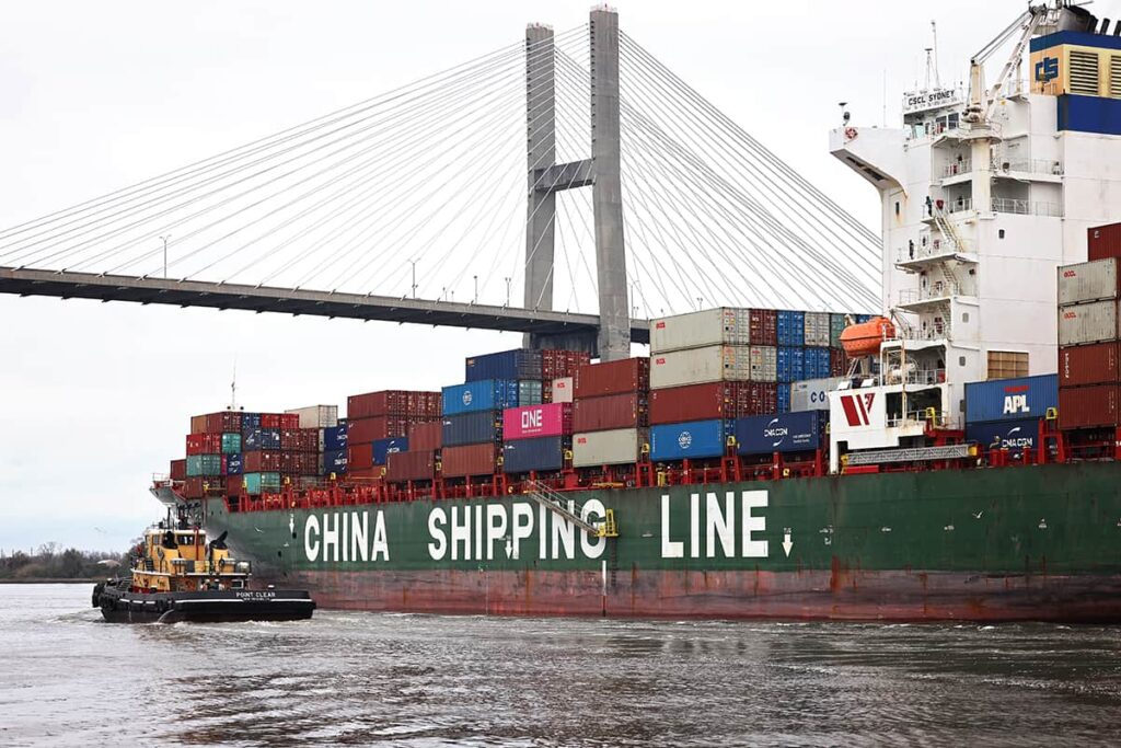A yellow tugboat leads a green cargo ship with under the Savannah bridge. The words CHINA SHIPPING LINE are visible on the cargo ship's port side