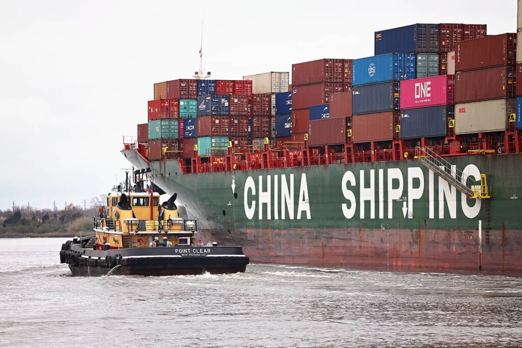 A yellow, red, and black tugboat hugs the port side of a cargo ship with the words CHINA SHIPPING on the side in chunky white lettering as they both head into the port of Savannah