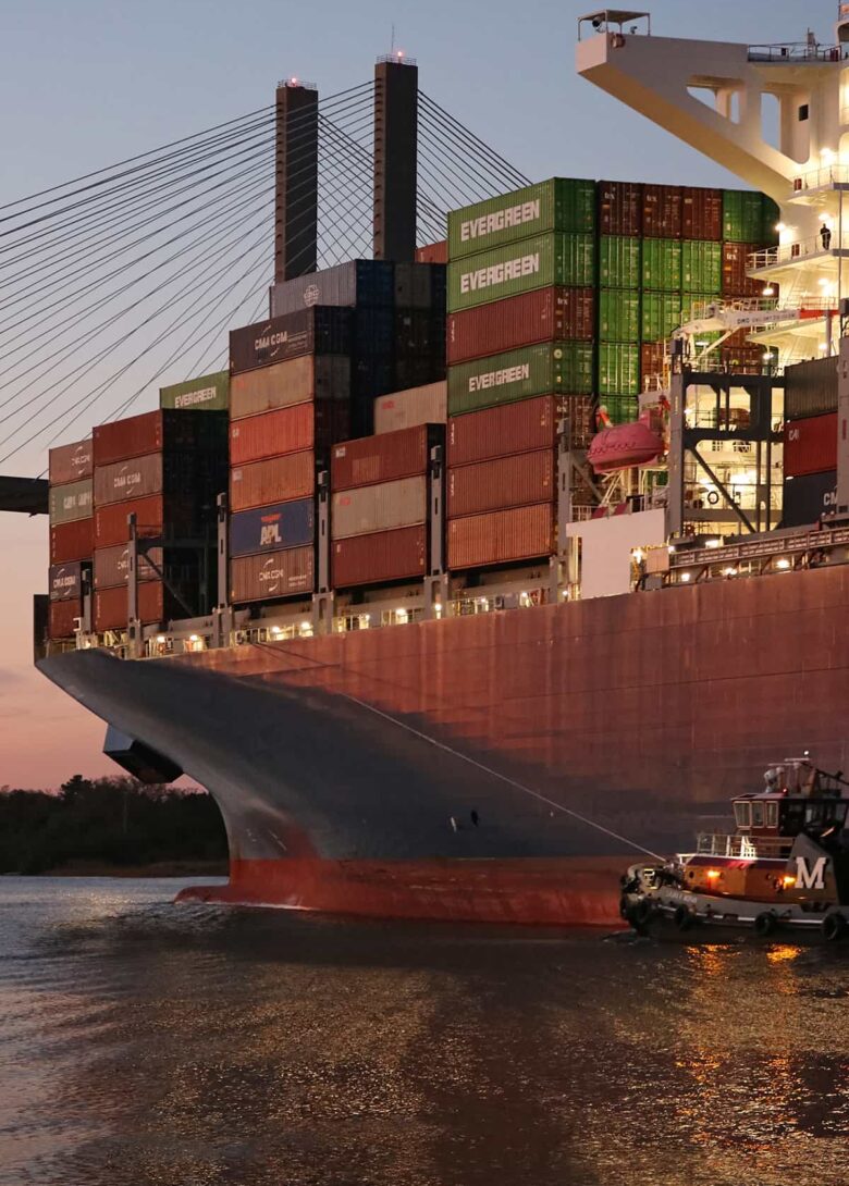 A massive cargo ship overshadows the Talmadge bridge at sunset along River Street in Savannah