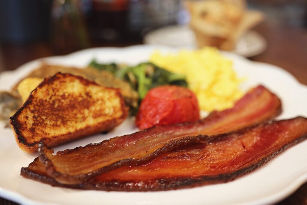 Close-up of a breakfast plate at Dottie's Market with two very thick slices of perfectly-cooked bacon in the foreground, and scrambled eggs, a cooked tomato, cornbread, hash browns, and greens slightly blurred in the background