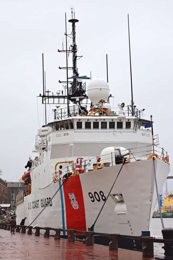 Bow of the USCGC TAHOMA docked along River Street. The cutter is white with one thin blue vertical stripe and one thick red vertical stripe on the side. The top deck is loaded with radar equipment. Black lettering on the side reads U.S. Coast Guard 908